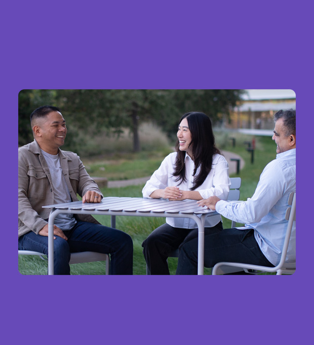 Three people of varying ages and ethnicities sit around a table conversing.